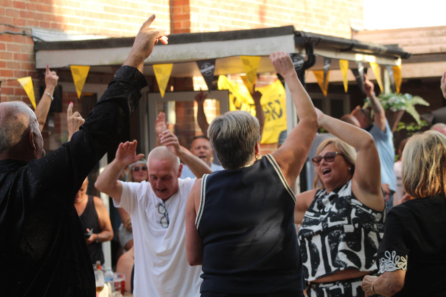 A group of people dancing and celebrating outdoors in front of a brick building, with yellow bunting flags hanging above them.